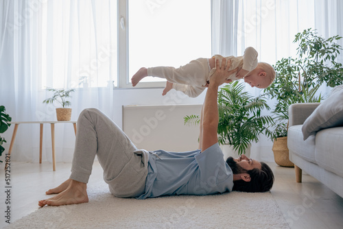 Side view of the young father enjoying time with his newborn baby, while holding him in air and looking at him with pleasure during playing at home. Father day concept