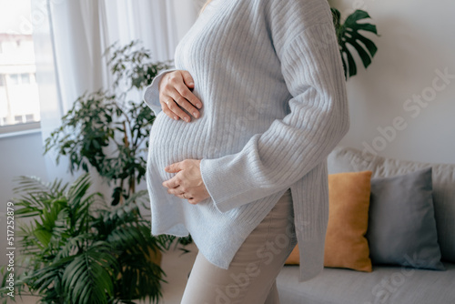 Close up view attractive pregnant woman is standing at the living room and holding her belly. Last months of pregnancy concept. Stock photo. Mothers Day