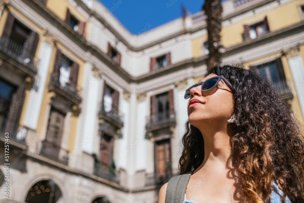 young dark and curly haired woman wearing sunglasses looking away. She has a blurred background of old buildings.