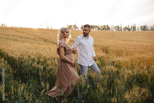 A pregnant woman in a beautiful long dress is walking through the field with her husband in a wheat field at sunset. Future parents.