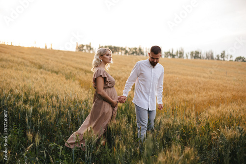 A pregnant woman in a beautiful long dress is walking through the field with husband in a wheat field at sunset. Future parents.