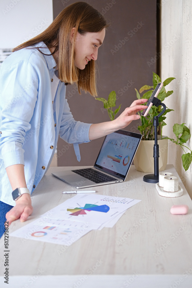 Joyful young woman holding graphs and charts and leading business meeting through video call. Joyful young woman holding graphs and charts and leading business meeting through video call.