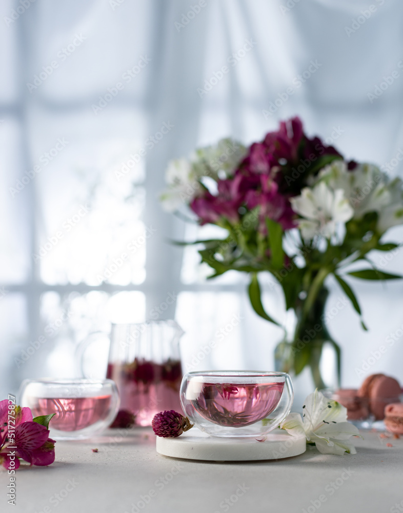 Pouring herbal or flower tea from glass teapot in a cup with macarons and flowers on white table in front of the window.Healthy hot drink in freeze motion.