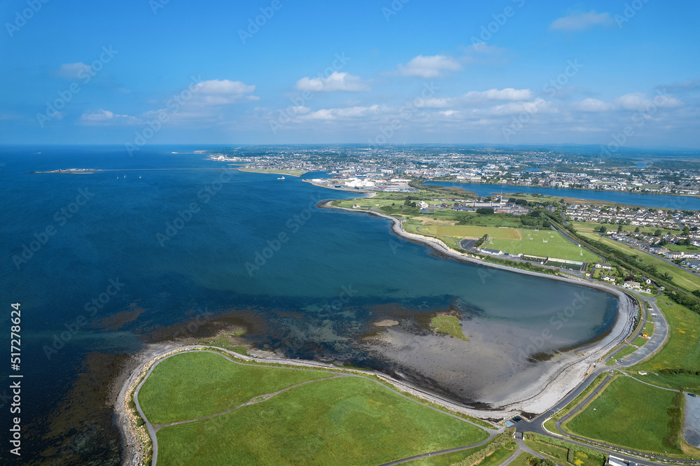 Aerial view on Galway city and Ballyloughane Strand. Warm sunny day. Blue sky and water of the ocean. High tide. Residential area and sport ground. Popular rest area with foot path for walk.