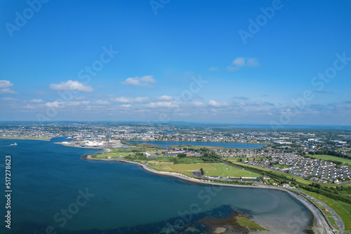 Aerial view on Galway city and Ballyloughane Strand. Warm sunny day. Blue sky and water of the ocean. High tide. Residential area and sport ground. Popular rest area with foot path for walk.