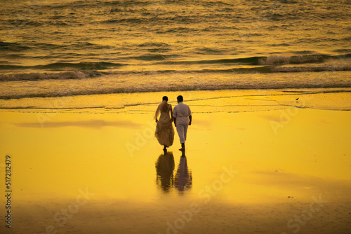 Loving couple walks along the beach after getting married in the golden sunlight. A Warm sea breeze fills the air while the orange sun reflects on the ocean making a perfect romantic summer evening photo