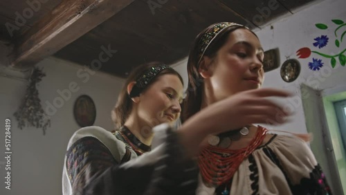 Young woman helps girl friend to put on red necklace closeup. Young woman helps girl friend to put on red necklace in old house. Stylish ladies wearing Ukrainian national outfit looks at each other photo