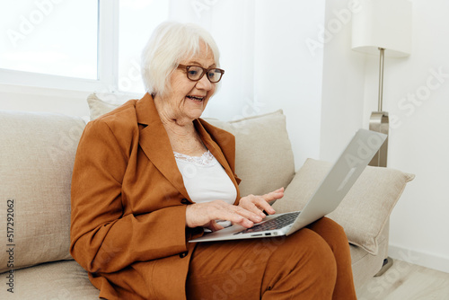 a pleasant, wise elderly woman is sitting looking at a laptop on the sofa in a relaxed pose in a bright room near the window with glasses on her face