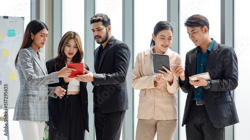 Group of millennial Asian Indian male female businessman businesswoman in formal suit standing holding tablet computer discussing sharing business ideas in multinational company office meeting room