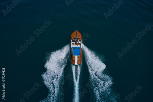 Wooden expensive italian boat with people fast movement on the water top view. High speed open modern wooden boat moving fast on dark water aerial view.