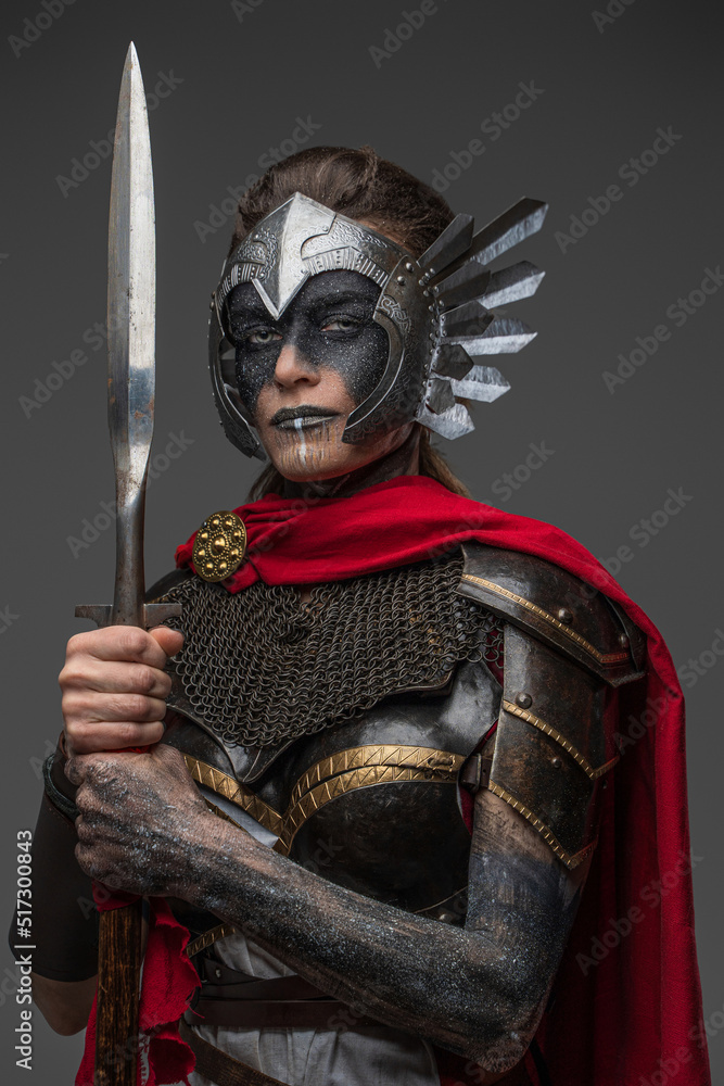 Fototapeta premium Shot of ancient valkyrie with make up dressed in dark armor with red cape holding spear.