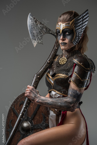 Принти на полотні Portrait of wild valkyrie dressed in armor holding huge axe and looking at camera against grey background