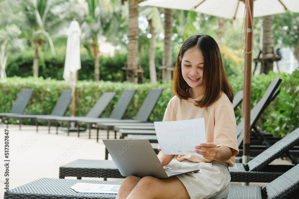 close-up A woman working at the swimming pool with her laptop computer holding paperwork in her hand. It is imperative for young Asian female tourists to sit and work on vacations at the hotel. close-up A woman working at the swimming pool with her laptop computer holding paperwork in her hand. It is imperative for young Asian female tourists to sit and work on vacations at the hotel.