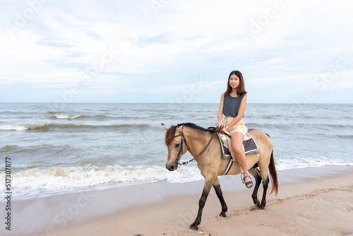 A young Asian woman is walking happily on a horse on the sandy beach before the sun goes down. Tourists come to relax at the beach, ride horses, and take a walk.