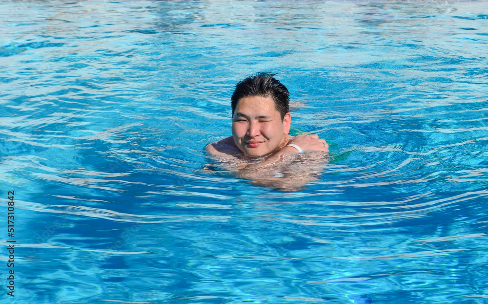 Asian man in the swimming pool. A young man swimming in the pool and holding a ball. Active resting. Asian male squinting eyes from the sun.