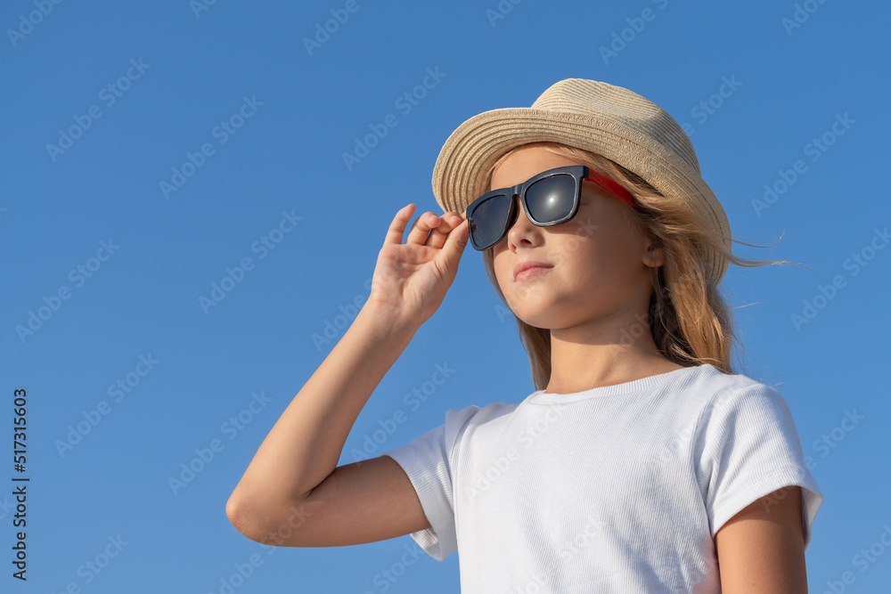 A young girl in a hat and sunglasses adjusts her glasses with one hand. A young girl in a hat and sunglasses adjusts her glasses with one hand.