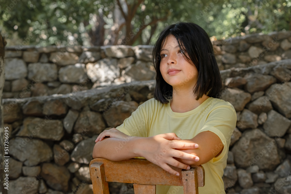 Fototapeta premium A young teenager girl sits on a wooden chair against a stone wall.
