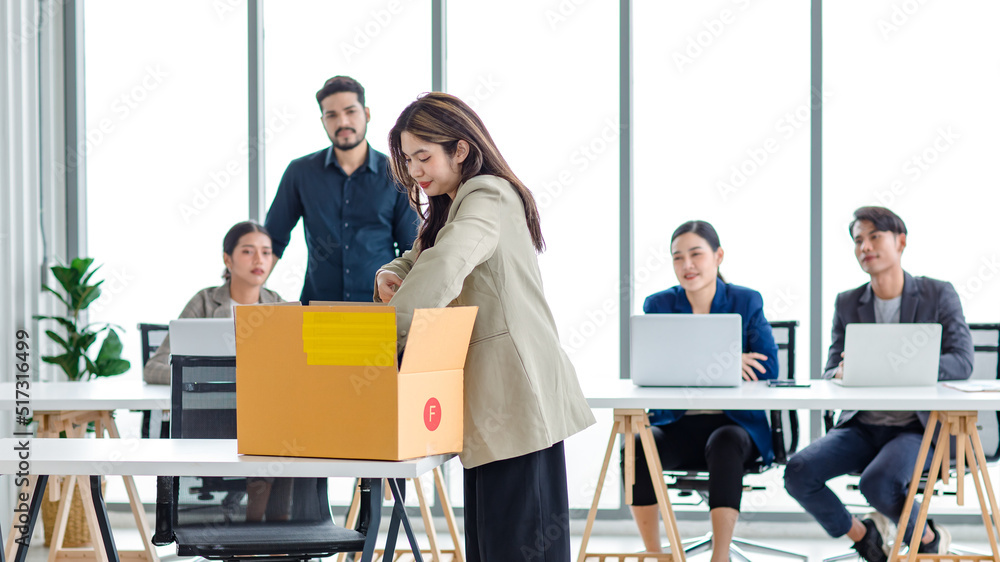 Fototapeta premium Portrait shot of Asian sad jobless businesswoman in casual suit standing holding belongings in cardboard box after fired while male and female colleagues waving hands goodbye in blurred background