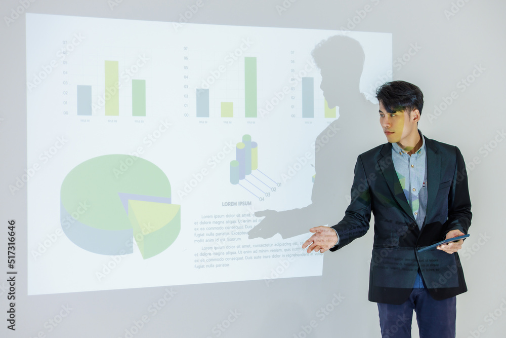 Millennial Asian young cheerful male businessman presenter in formal suit standing holding notebook and pen pointing presenting corporate graph chart data information from wall screen in meeting room. - obrazy, fototapety, plakaty 