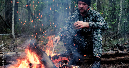 Man with cup of tea the fire in the forest. Human looks at the bonfire and drinks tea on the nature in the summer. Alone traveler sits and rests by the fire with a cup of coffee at dusk.