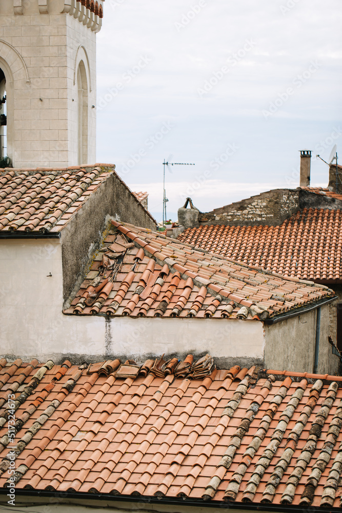 Antique pitched roof with old red tiles on an Italian house. In the background are a white tower, red roof, and brick chimneys.