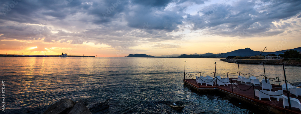 Naklejka premium Horizontal image of the view of a bay at sunrise. Jetty with chairs and a boat skyline in the background.