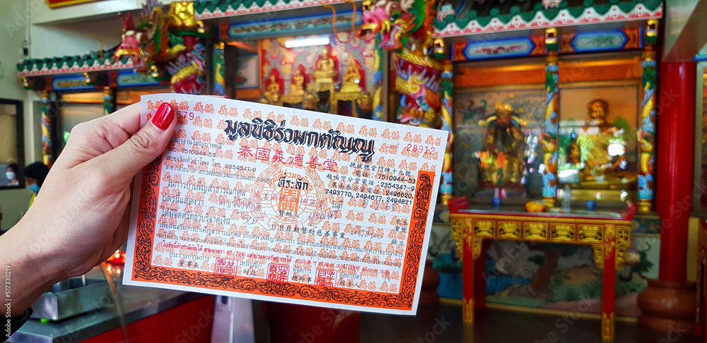 Naklejka premium ฺBangkok, Thailand - July 1, 2022: Holding receipt of funeral donation after paying merit to a corpse without relatives with Chinese god statue and copy space at China temple or wat hua lampong.