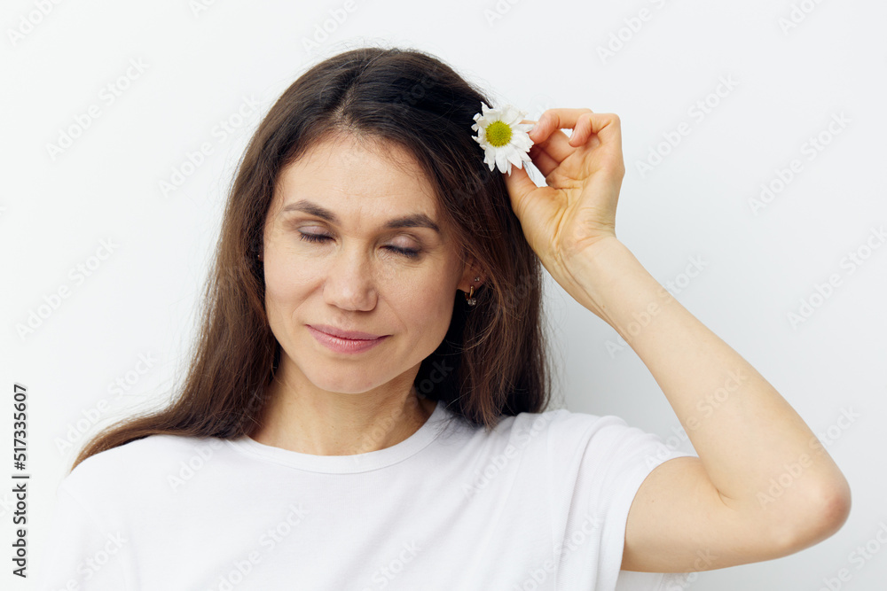 portrait of a cute, happy, beautiful brunette woman, in a white tank top, smiling pleasantly, inserting a chamomile flower into her hair with her eyes closed portrait of a cute, happy, beautiful brunette woman, in a white tank top, smiling pleasantly, inserting a chamomile flower into her hair with her eyes closed