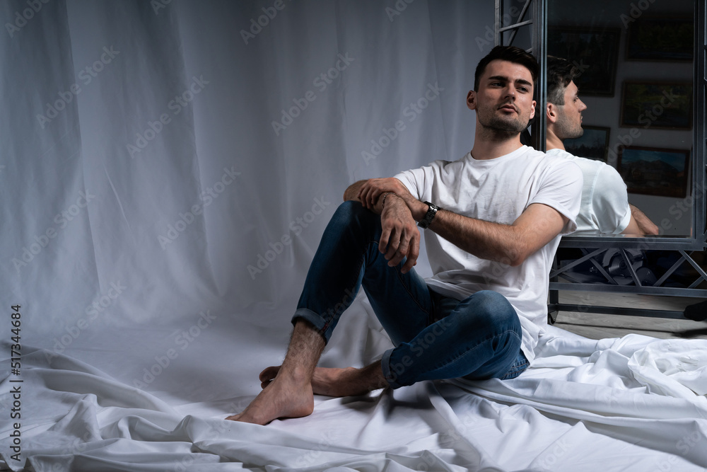 male portrait. the guy in the studio sits near the mirror. reflection of a man in the mirror. twilight. brutal image. jeans, white t-shirt. focus on the person