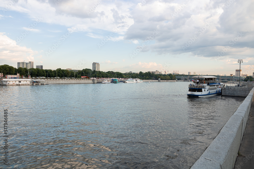 Naklejka premium Moscow river with pleasure crafts. Russian flag on one of the boats. Park with trees along the waterfront. Houses and buildings in the distance.