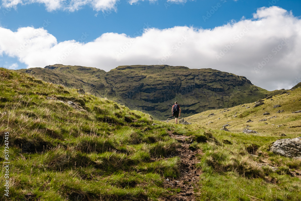 Naklejka premium A hillwalker hiking up a hill trail with a view of a mountain. Beautiful green Scottish mountain scenery in the Trossachs National Park, summer