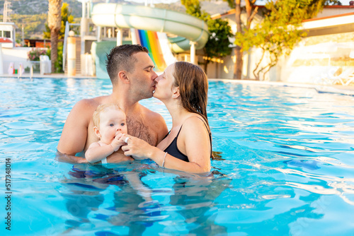 happy family mom, dad and baby daughter are swimming in the pool with water slides and having fun on vacation, kissing and hugging