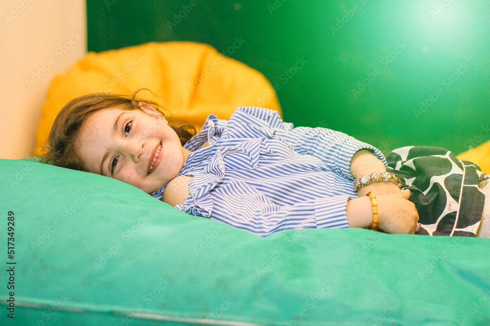 Happy smiling girl lying on green mattress in kindergarten with multi colored interior and looking at camera carefully