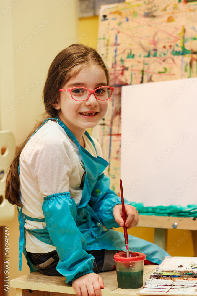 Little painter girl with eyeglasses sitting in front of the easel, holding the brush and ready to paint something