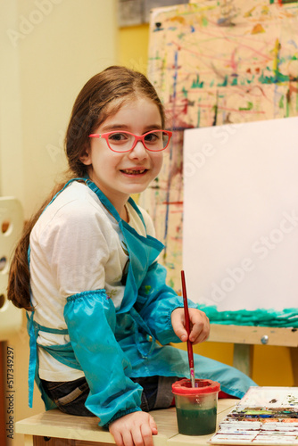 Obraz na płótnie Little painter girl with eyeglasses sitting in front of the easel, holding the brush and ready to paint something w salonie