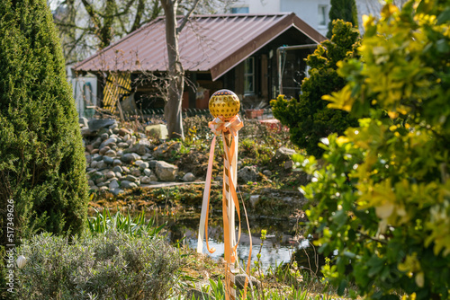 homemade Easter decor in the form of a pole with a glass ball and ribbons installed in the garden near a private house
