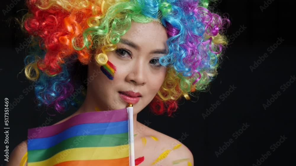 Slow motion, portrait of young woman in rainbow wig with paint on her cheeks and holding a rainbow flag on black background. High quality 4k footage Slow motion, portrait of young woman in rainbow wig with paint on her cheeks and holding a rainbow flag on black background. High quality 4k footage
