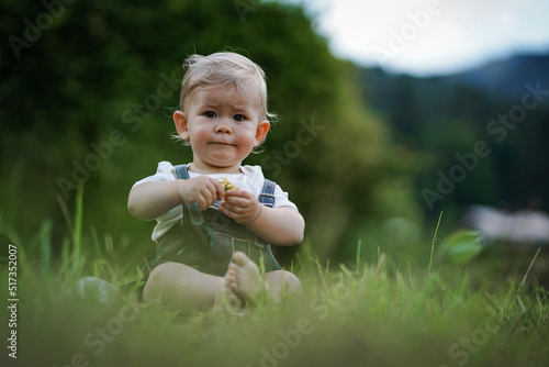 one year old blond german baby boy in bavarian dress with lederhose sitting outside on the meadow with a yellow flower to congratulate for birthday photo