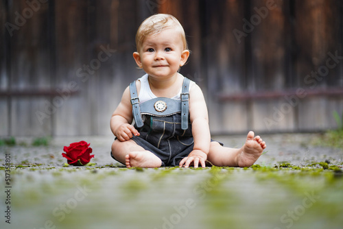 happy smiling one year old baby boy sitting or crawling in bavarian lether pants called Lederhosn outdoor on the floor with a red rose to congratulate for birthday
 photo