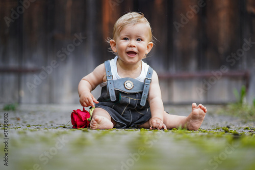 happy smiling one year old baby boy sitting or crawling in bavarian lether pants called Lederhosn outdoor on the floor with a red rose to congratulate for birthday 