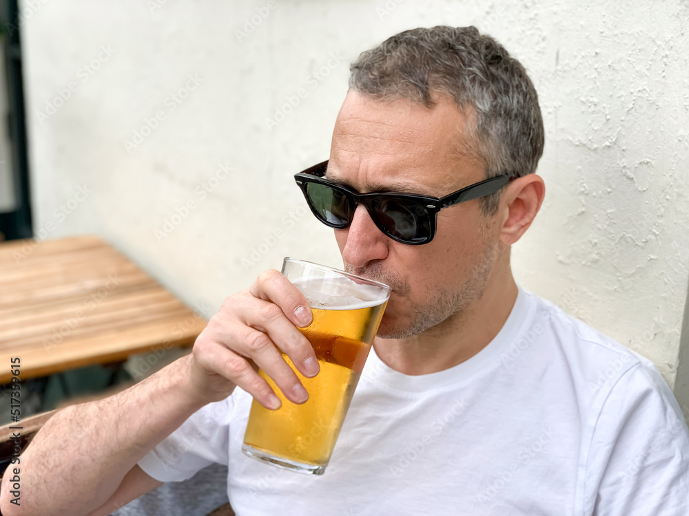 Middle aged man is drinking beer in a cafe or restaurant pub outdoor. Drinking alcohol. Candid. Refreshment, summer time, pub garden. wearing white t-shirt and sunglasses. Drinking lager or ale beer. Middle aged man is drinking beer in a cafe or restaurant pub outdoor. Drinking alcohol. Candid. Refreshment, summer time, pub garden. wearing white t-shirt and sunglasses. Drinking lager or ale beer.