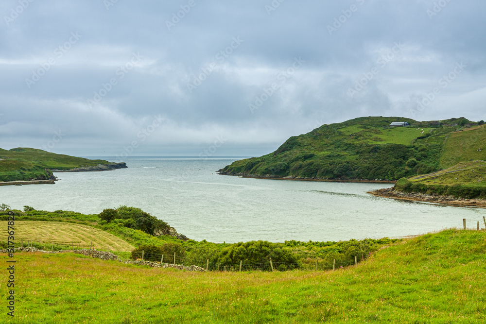 A wide bay with hills covered with green trees under a thick cloudy sky. The sandy beach of a cove-shaped bay with lush green grass near the edge of the ocean.