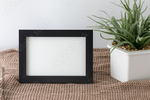 Blank horizontal black frame beside a houseplant, painting or artwork display on brown cloth