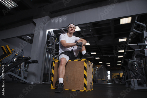 Low angle full length shot of a handsome athletic man sitting on a jumping box at gym, resting after exercising
