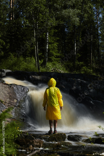 Concept of traveling in Russia in summer. Female tourist in stylish yellow raincoat stands near waterfall and enjoys splashing cold water on hot summer day. Waterfall of Karelia. Rear view.