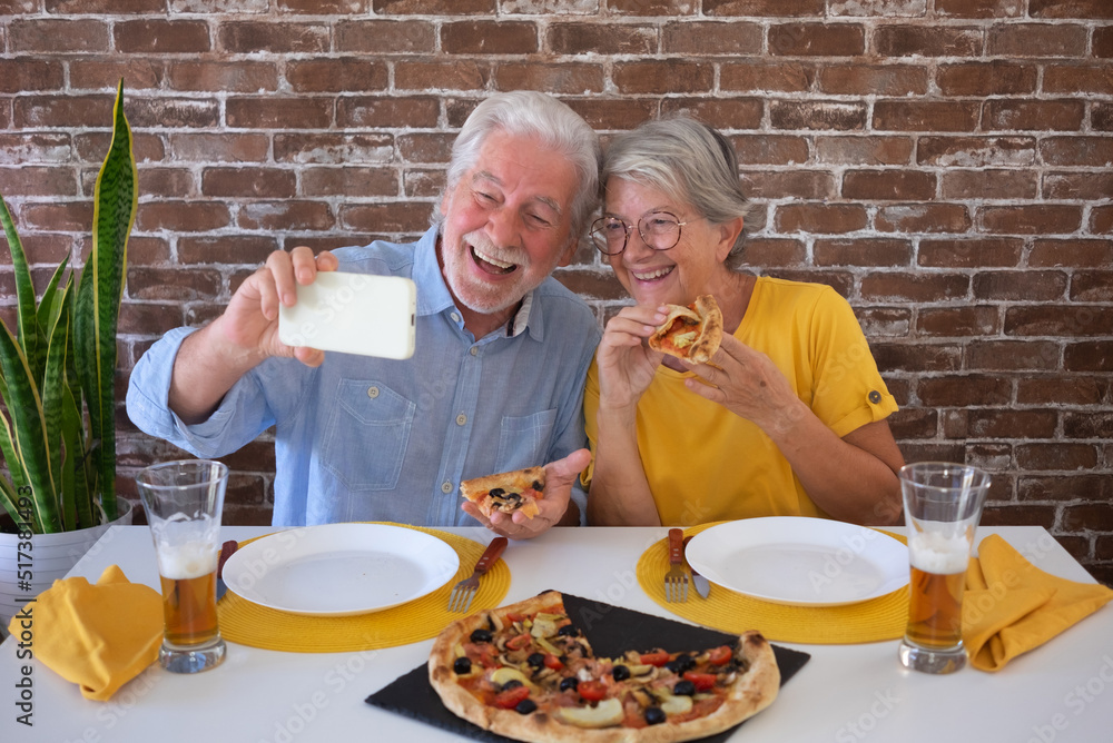 Cheerful senior couple having fun together eating a pizza while taking selfie with smartphone. Sitting at home table with beer and pizza. Celebrating holiday in company Cheerful senior couple having fun together eating a pizza while taking selfie with smartphone. Sitting at home table with beer and pizza. Celebrating holiday in company
