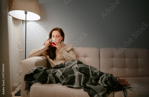 Young woman caught a cold by wrapping herself in a blanket at home and drinking hot tea from a cup.