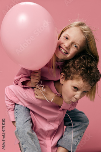 joyful, happy children of school age, on a pink background in pink clothes play and the boy rolls the girl with a balloon on her back