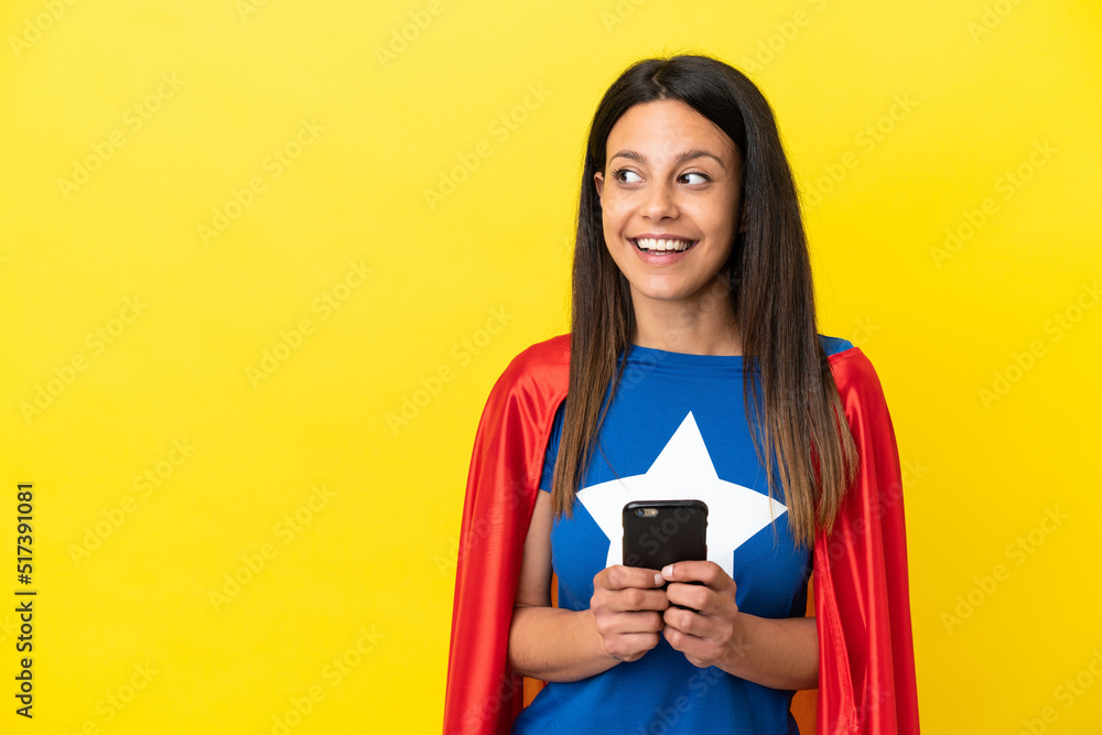 Super Hero woman isolated on yellow background using mobile phone and looking up Super Hero woman isolated on yellow background using mobile phone and looking up