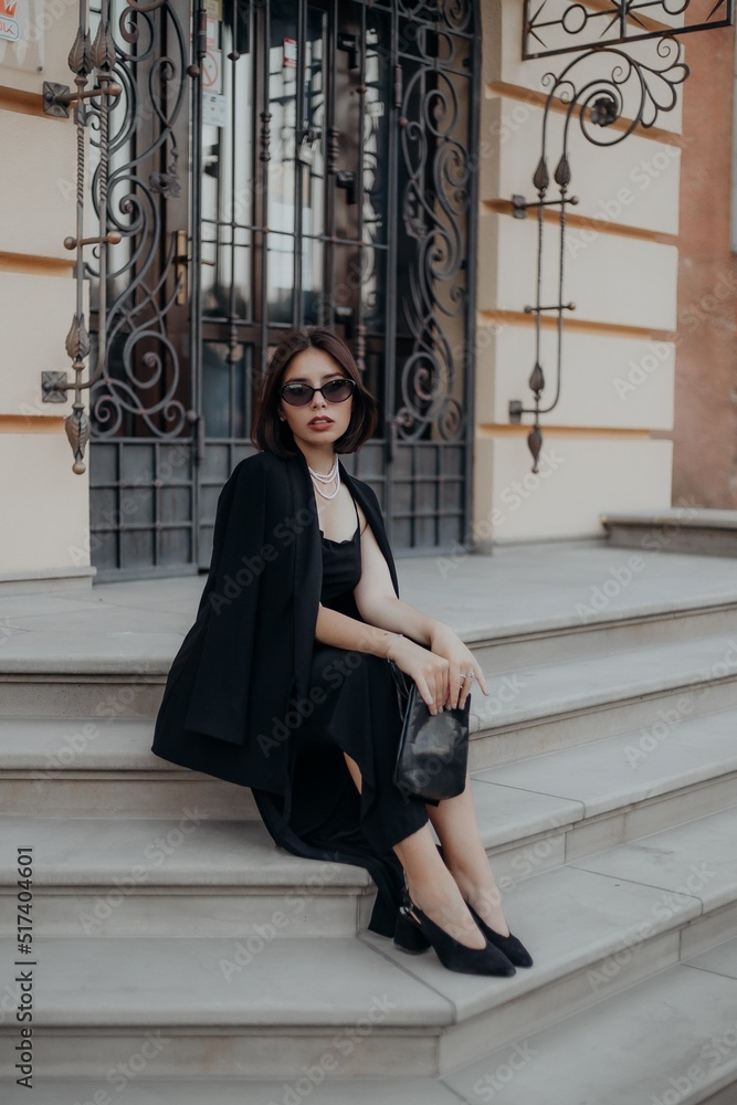 a brunette girl in a black jacket and dress and black glasses sits in the city with a handbag a brunette girl in a black jacket and dress and black glasses sits in the city with a handbag
